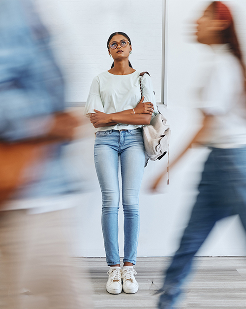 A young woman stands still against a blurred background of moving people, wearing a light-colored shirt and jeans, holding a backpack, and looking contemplative in a busy environment.